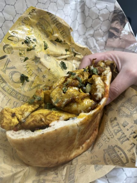 A hand holds a pita bread filled with grilled chicken, melted cheese, herbs, and sauce, wrapped in branded Altim Grill paper—fresh from the bakery.