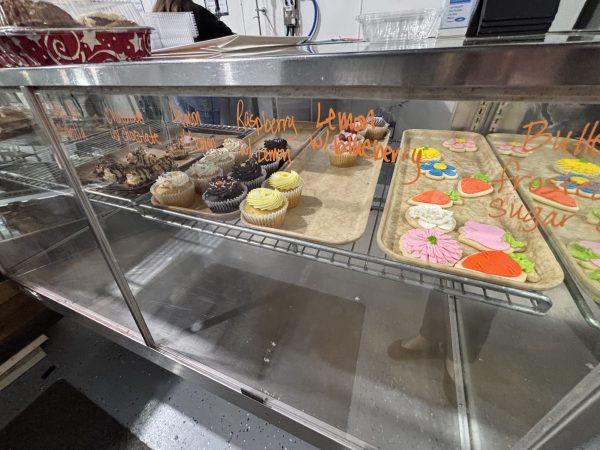 Display case with trays of assorted cupcakes and decorated sugar cookies, labeled with flavors like lemon, raspberry, chocolate, and buttercream.