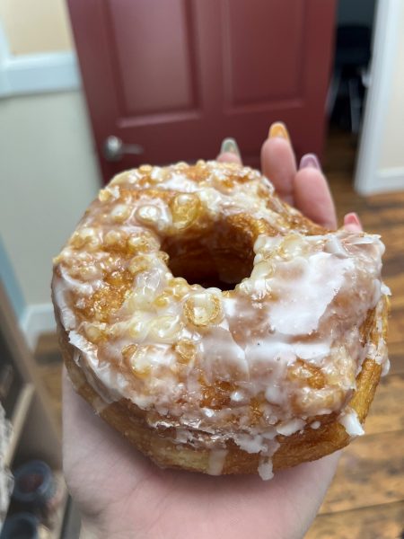 A hand holds a glazed cronut with visible icing and a textured, flaky surface. A red door and wooden floor are in the background.