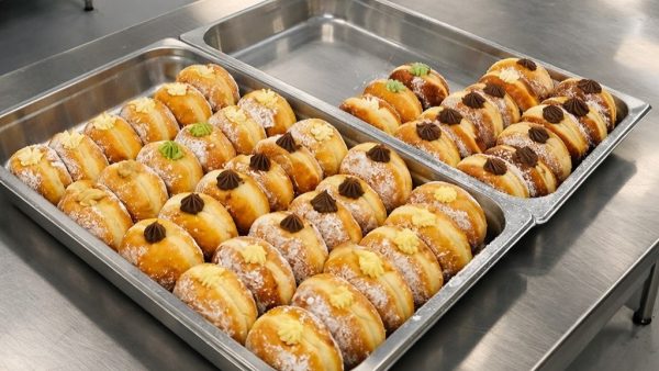 Two metal trays filled with rows of round, sugar-dusted donuts, each topped with a dollop of cream or chocolate and garnished with small decorations.
