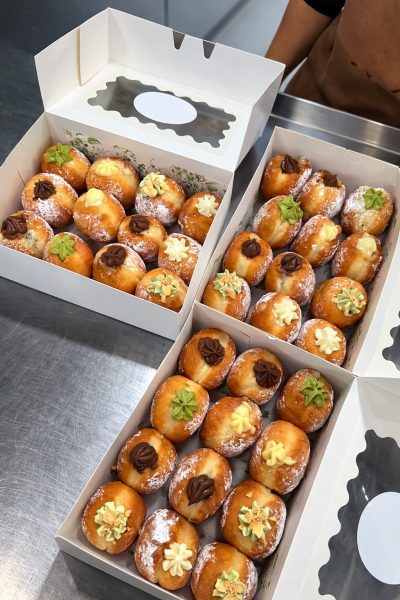 Three boxes of round, filled doughnuts topped with various cream decorations are displayed on a stainless steel counter.