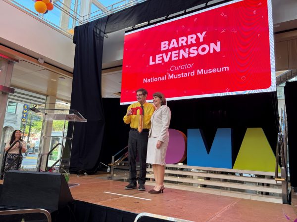 Barry Levenson and a woman stand on stage in front of a large screen reading “Barry Levenson, Curator, National Mustard Museum” at an indoor event.