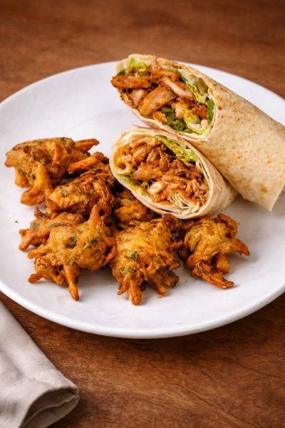 A white plate with a sliced chicken wrap filled with lettuce and several pieces of fried vegetable pakoras sits on a wooden cafe table.