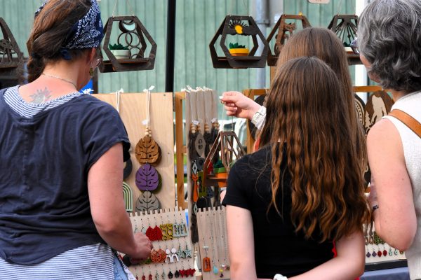 Four women browse handmade jewelry and wooden crafts displayed on a market stall, with colorful earrings and pendants hanging on boards.