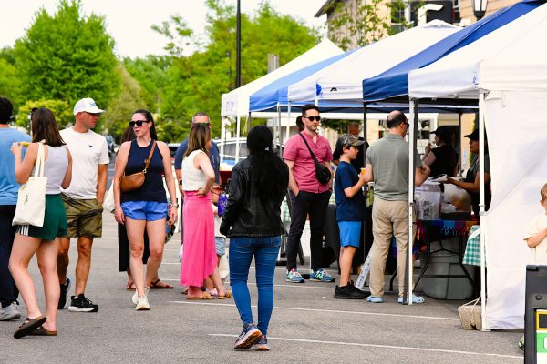 People walk and browse booths at an outdoor market or fair, with tents set up and trees in the background.