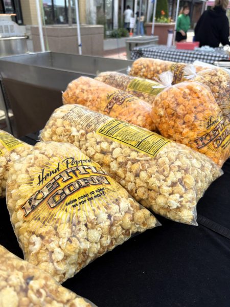 Bags of hand-popped kettle corn are displayed on a table at the Middleton Farmers Market, with vendors and shoppers in the background.