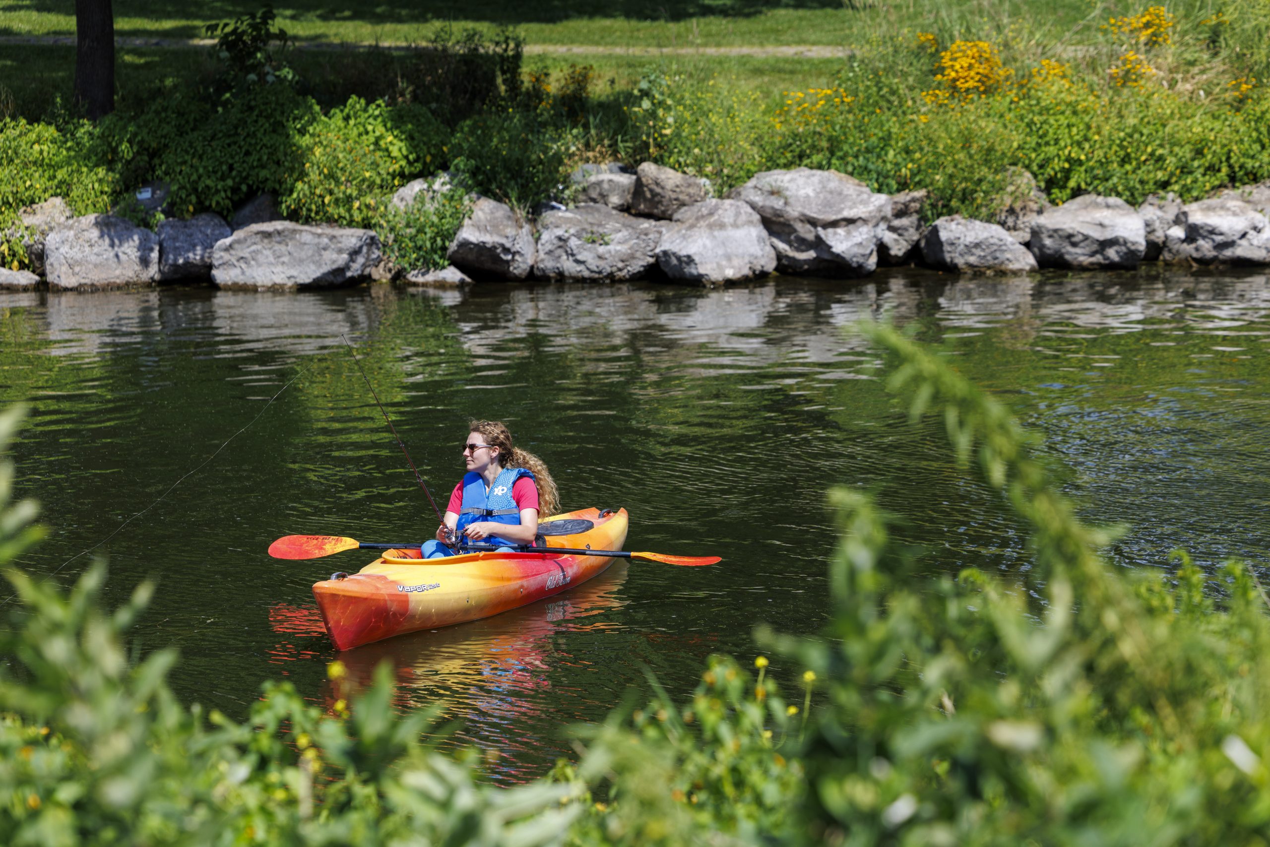 A person wearing a life jacket paddles a red and yellow kayak on a calm river, with rocks, green plants, and yellow flowers along the riverbank.