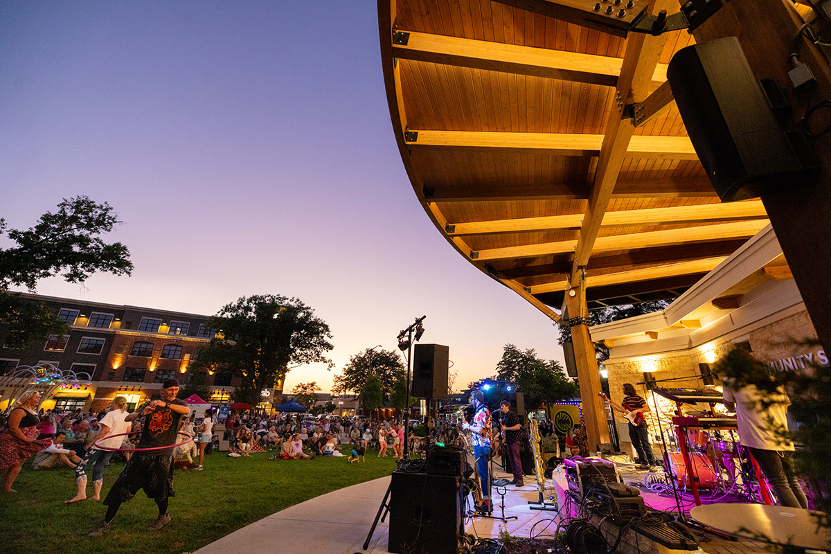 People gather on a lawn at sunset to watch a live band performing on an outdoor stage with large wooden beams.