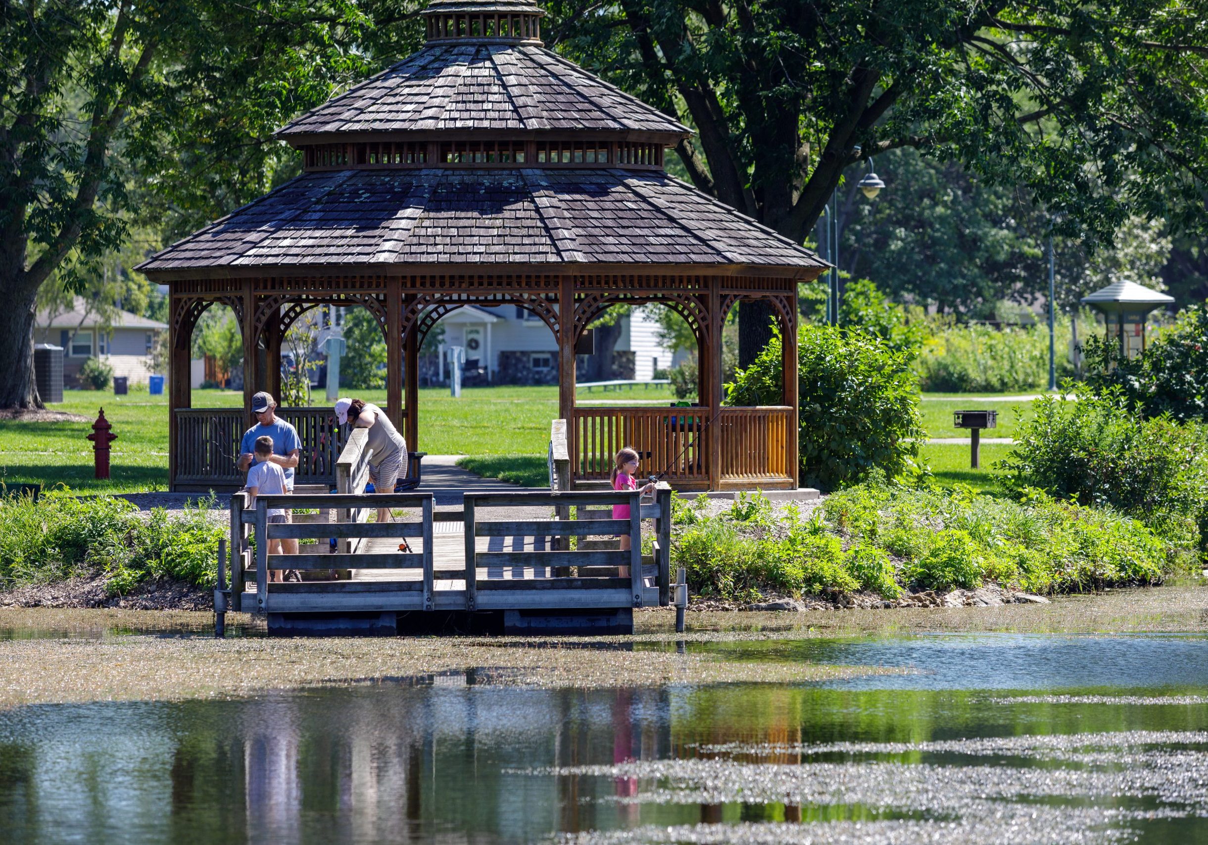 A group of people stands on a dock by a pond in front of a wooden gazebo in a green park on a sunny day.