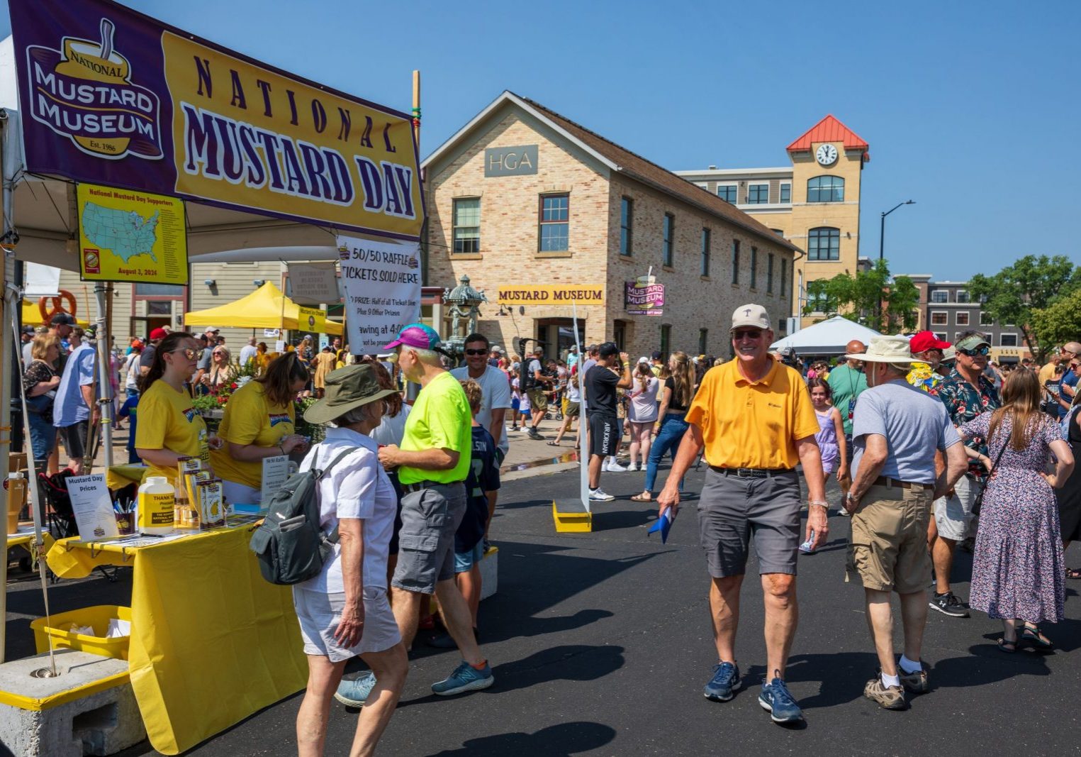 People attend an outdoor event for National Mustard Day, with a booth and signs for the Mustard Museum visible among the crowd on a sunny day.
