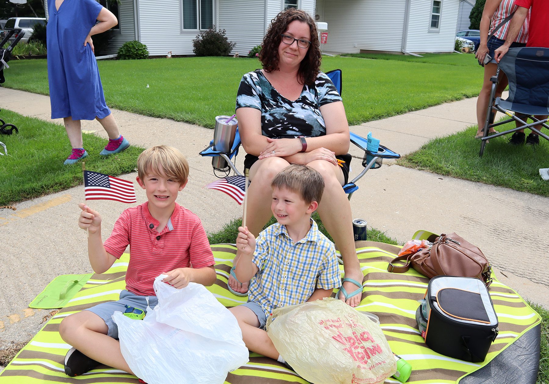 A woman sits on a folding chair behind two young boys holding small American flags, seated on a striped blanket with bags and a cooler beside them, all waiting for the good neighbor festival parade to begin.