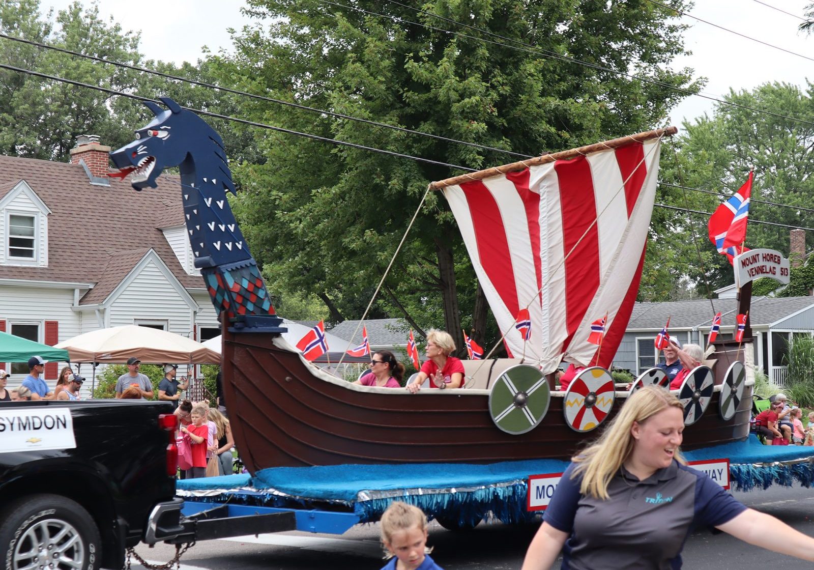 A parade float designed as a Viking ship with people on board, pulled by a vehicle, brings excitement to the Good Neighbor Festival as it passes by houses and spectators enjoying live music on a lively residential street.