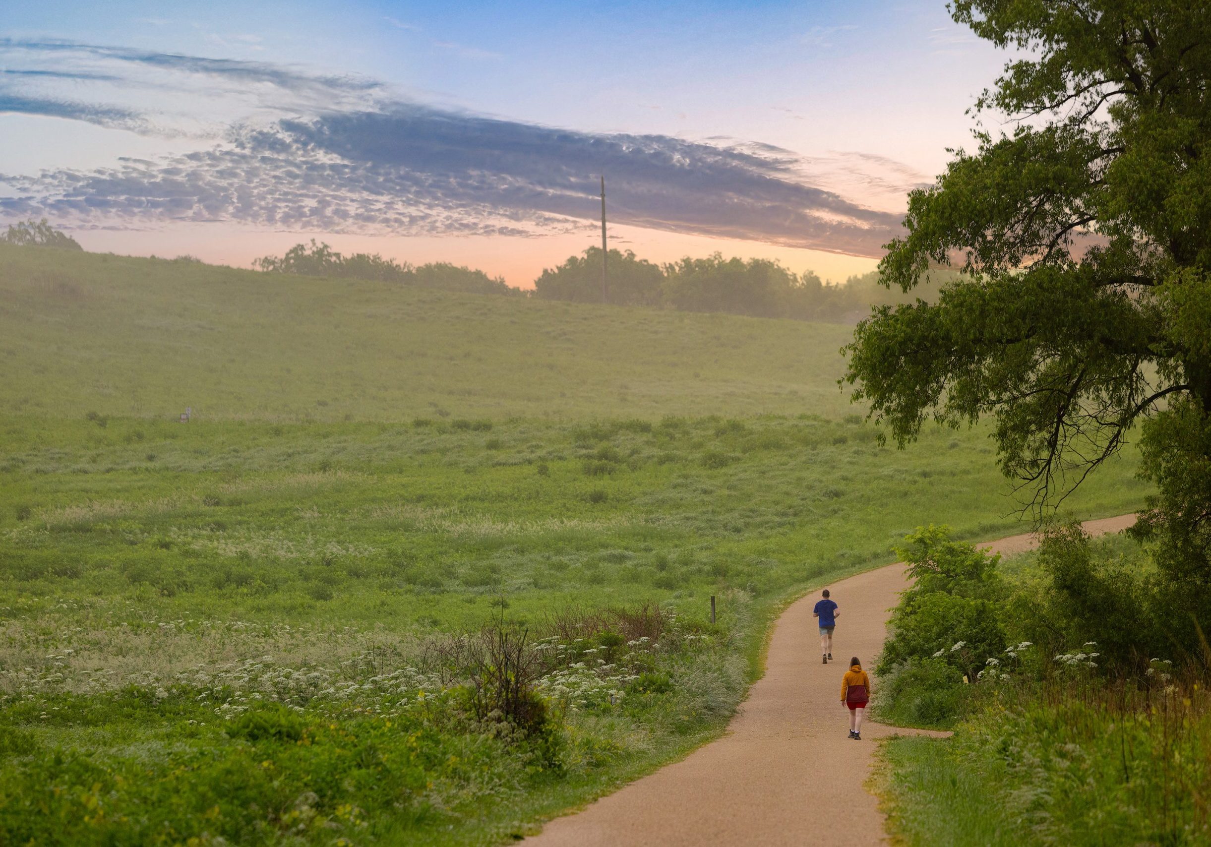 Two people walk along a winding trail through a green field on a golf course under a partly cloudy sky at sunrise or sunset.