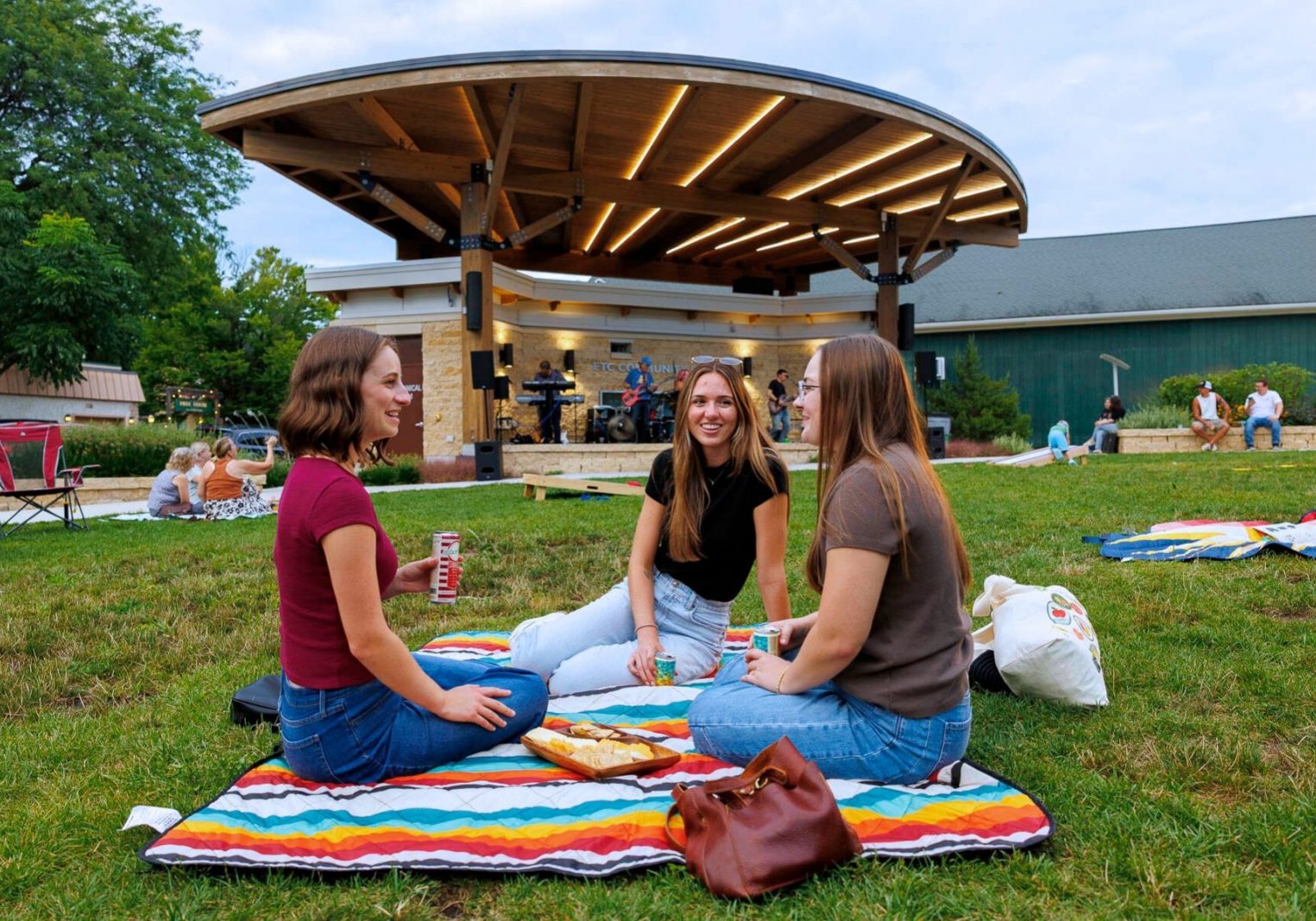 Three women sit on a blanket in a grassy area, talking and eating, with a band performing on a stage under a wooden pavilion in the background.